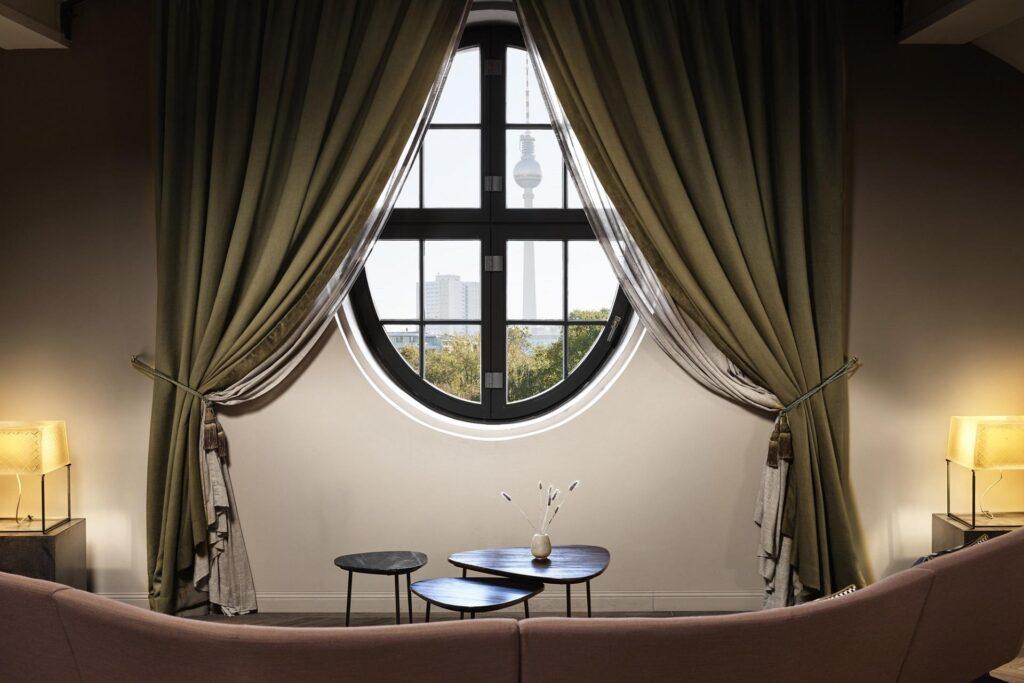 Ein rundes, von grünen Vorhängen umrahmtes Fenster gibt den Blick frei auf eine Stadtlandschaft mit einem Turm, hinter dem sich eine Sitzgruppe mit kleinen Tischen und dekorativen Vasen befindet, die von zwei Tischlampen beleuchtet wird.