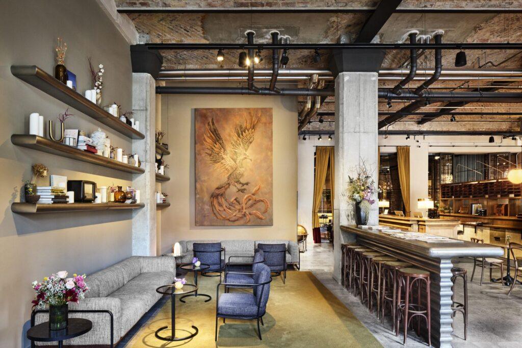 Moderner Lounge- und Barbereich mit freiliegender Decke, großem Kunstwerk eines Vogels, gepolsterten Sitzgelegenheiten, runden Tischen, Wandregalen und einem langen Tresen mit Barhockern.