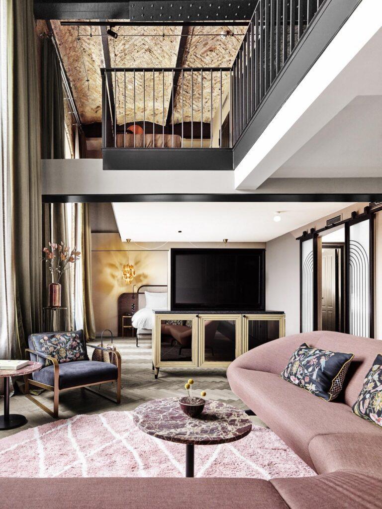 Ein moderner Wohnbereich im Loft-Stil mit einem rosafarbenen, geschwungenen Sofa, geblümten Sesseln, einem Marmor-Couchtisch und einem darüber liegenden Schlafzimmer im Zwischengeschoss.