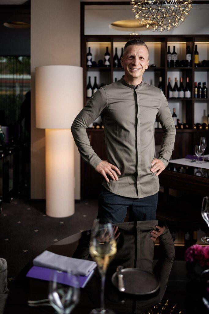 Ein Mann steht lächelnd mit in die Hüften gestemmten Händen in einem modernen Restaurant, mit Weinflaschen in Regalen hinter ihm und einem gedeckten Esstisch im Vordergrund.