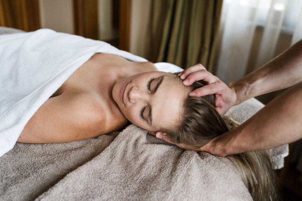 Eine Person, die auf einem Massagetisch liegt und ein Handtuch über sich drapiert hat, erhält eine Kopfmassage durch die Hände einer anderen Person.