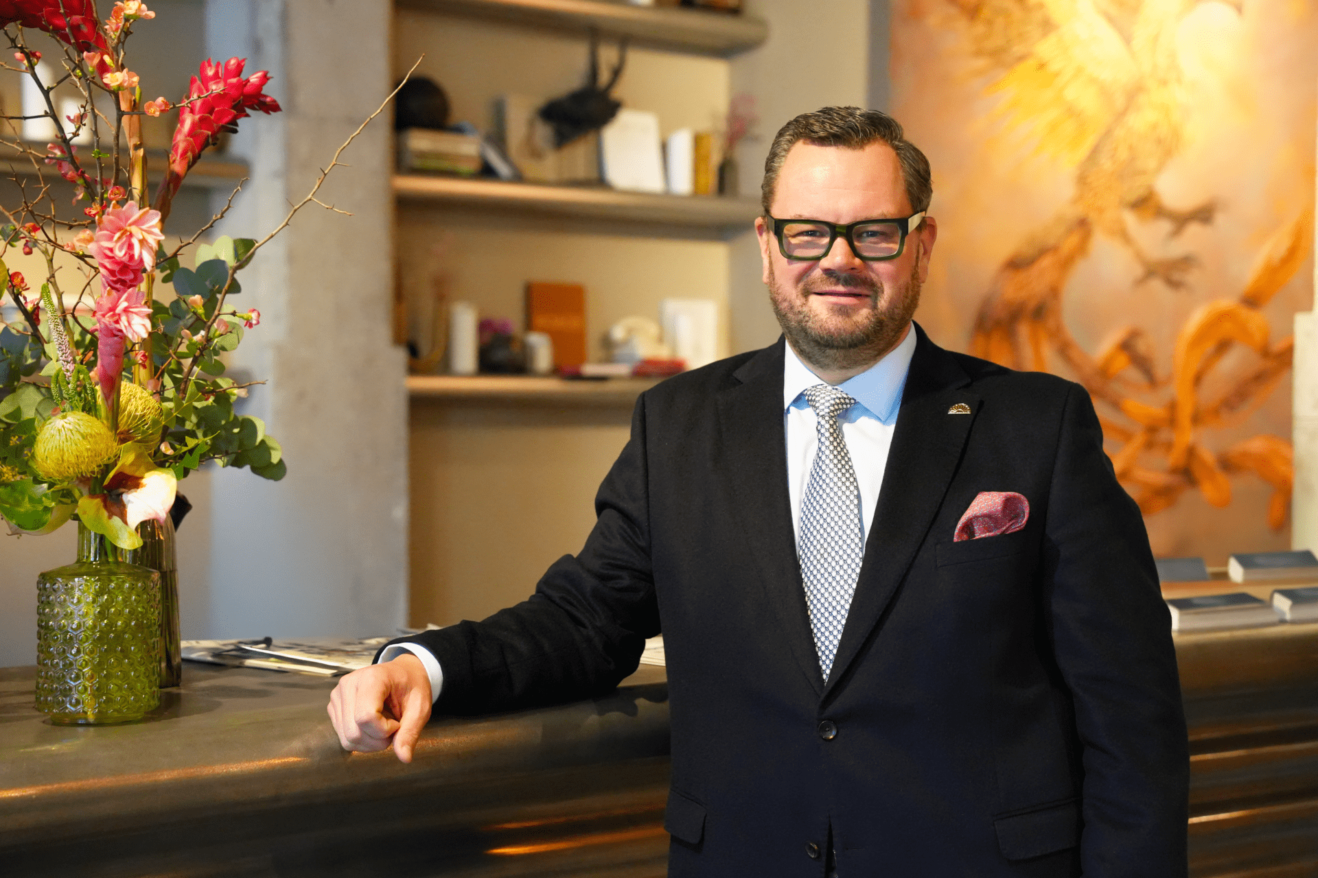 Ein Mann in Anzug und Krawatte steht an einem Tresen mit einer Blumenvase, Bücherregalen und Kunstwerken im Hintergrund.
