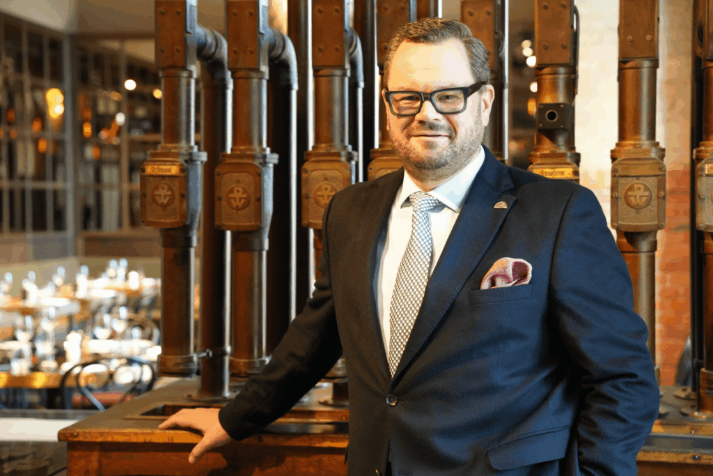 Ein Mann im Anzug und mit Brille steht in einem Innenraum und stützt sich auf eine hölzerne Fläche, im Hintergrund sind Industrierohre und ein Restaurant zu sehen.