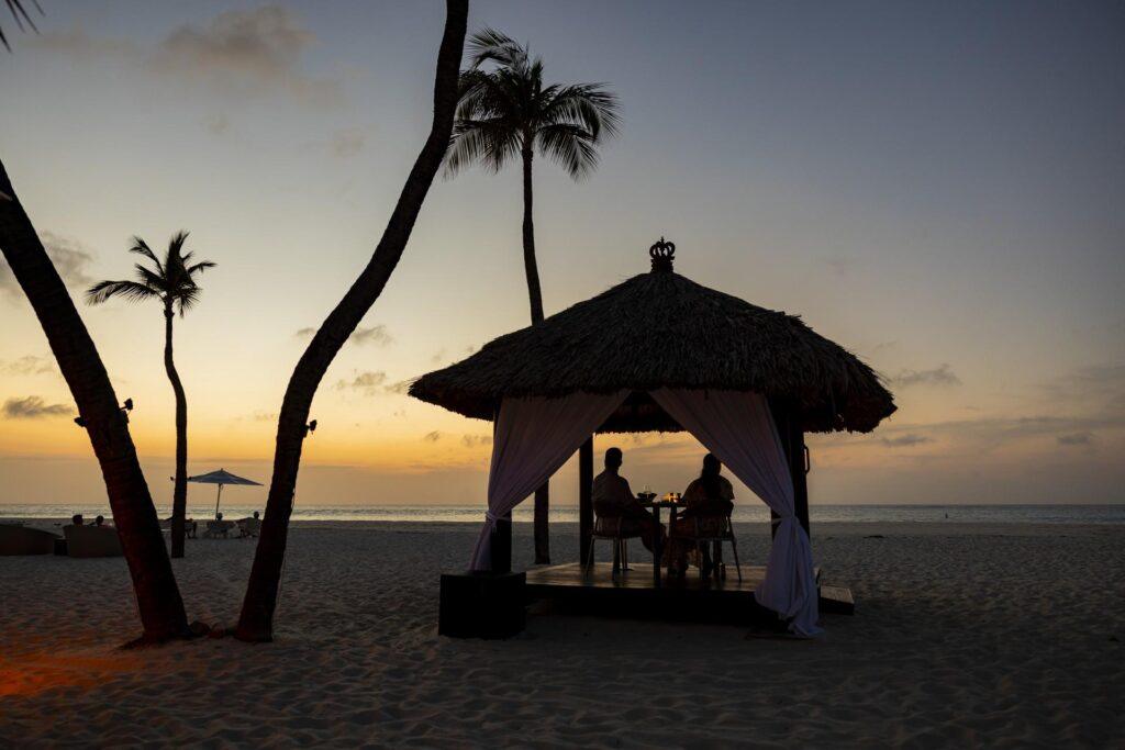 Zwei Personen sitzen an einem Tisch unter einer strohgedeckten Cabana an einem Sandstrand bei Sonnenuntergang, mit Palmen und dem Meer im Hintergrund.
