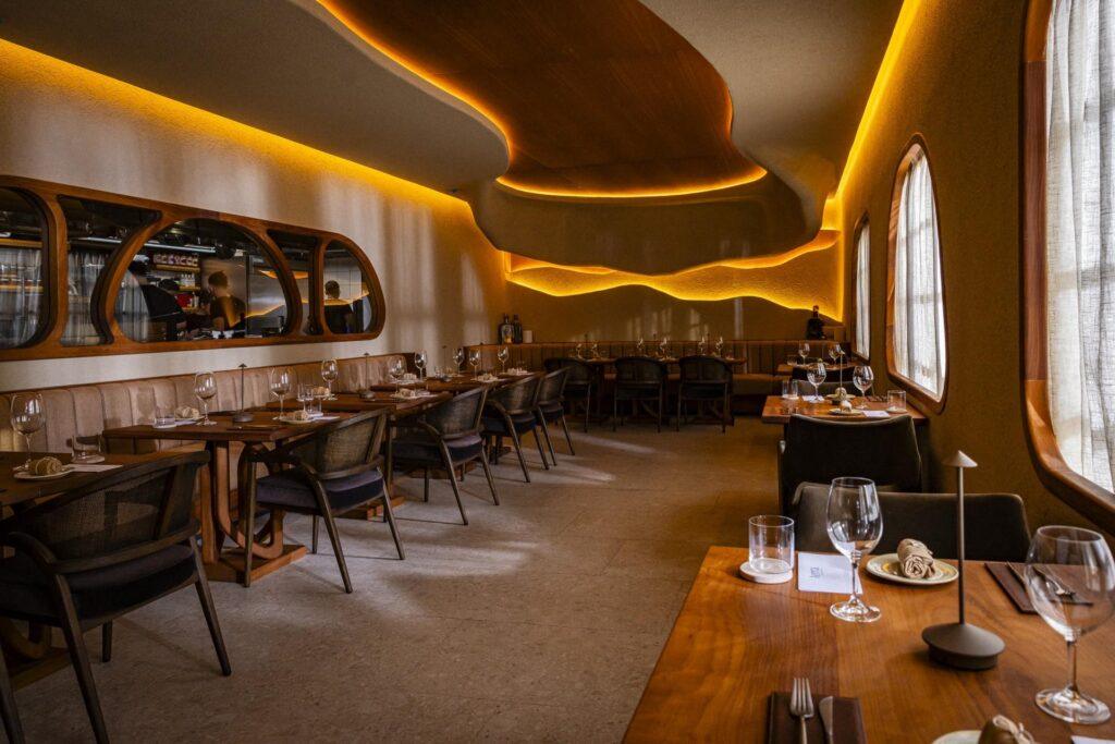 Ein modernes Restaurant mit Holztischen, gepolsterten Stühlen, Essgeschirr, geschwungenen Decken, warmer Beleuchtung und großen Fenstern mit transparenten Vorhängen.